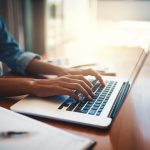 Adult sitting at a table typing on a laptop.